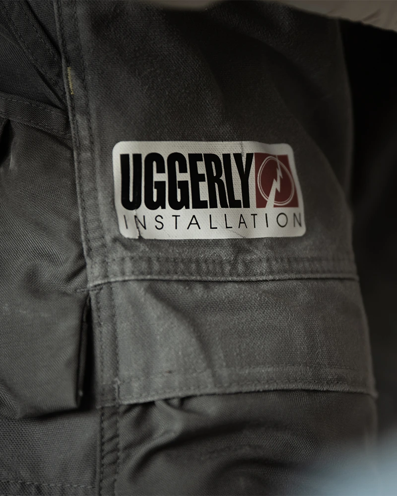Et nærbillede af et par grå arbejdsbukser, hvor man kan et logo af Uggerly Installation.